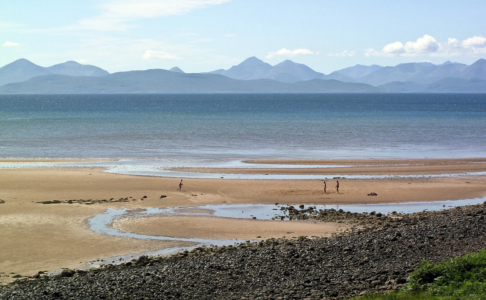Zand en stenen langs Applecross Bay, met enkele wandelaars en bergsilhouetten aan de overkant van het water.