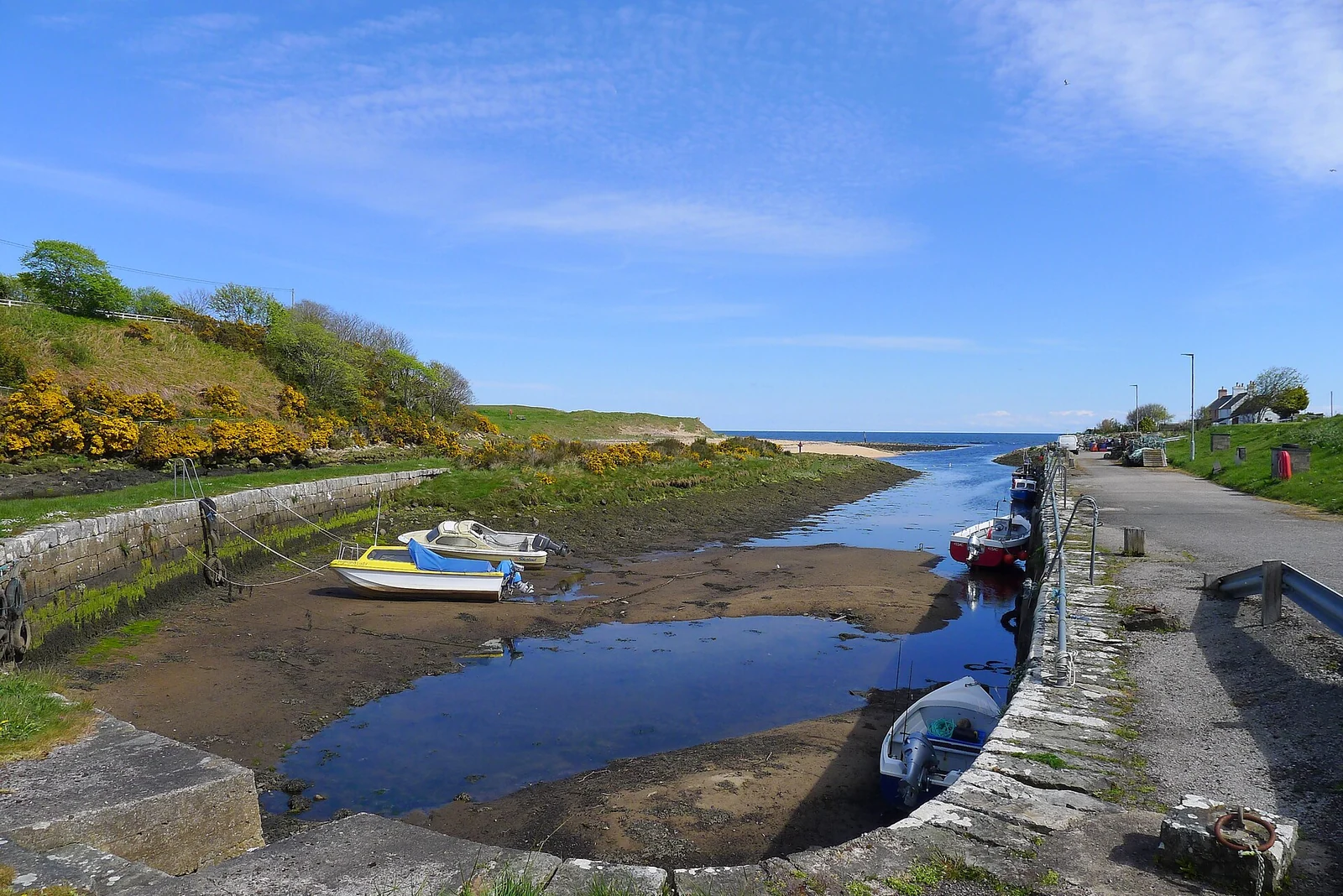 Kleine boten liggen bij laag water in de smalle haven van Brora, tussen stenen kades met de zee in de verte.
