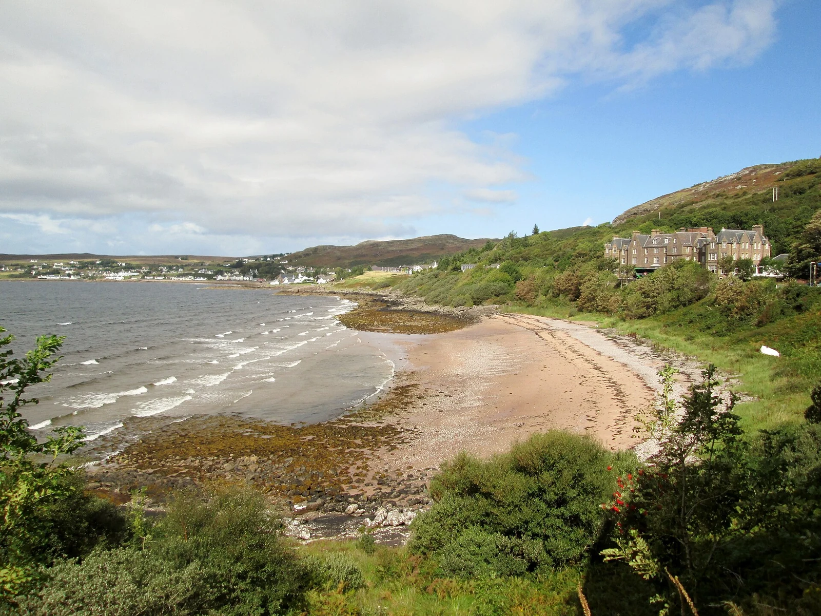 Een gebogen zandstrand bij Gairloch met kleine golven, groene struiken en bebouwing langs de baai.