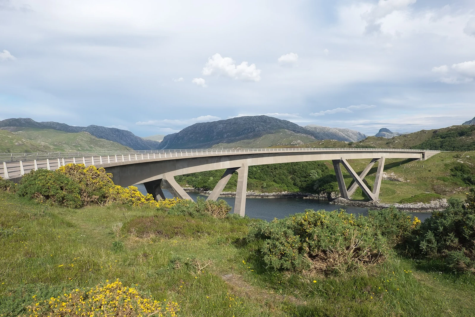 De gebogen betonnen brug bij Kylesku overspant het water tussen groene oevers en afgeronde bergen.