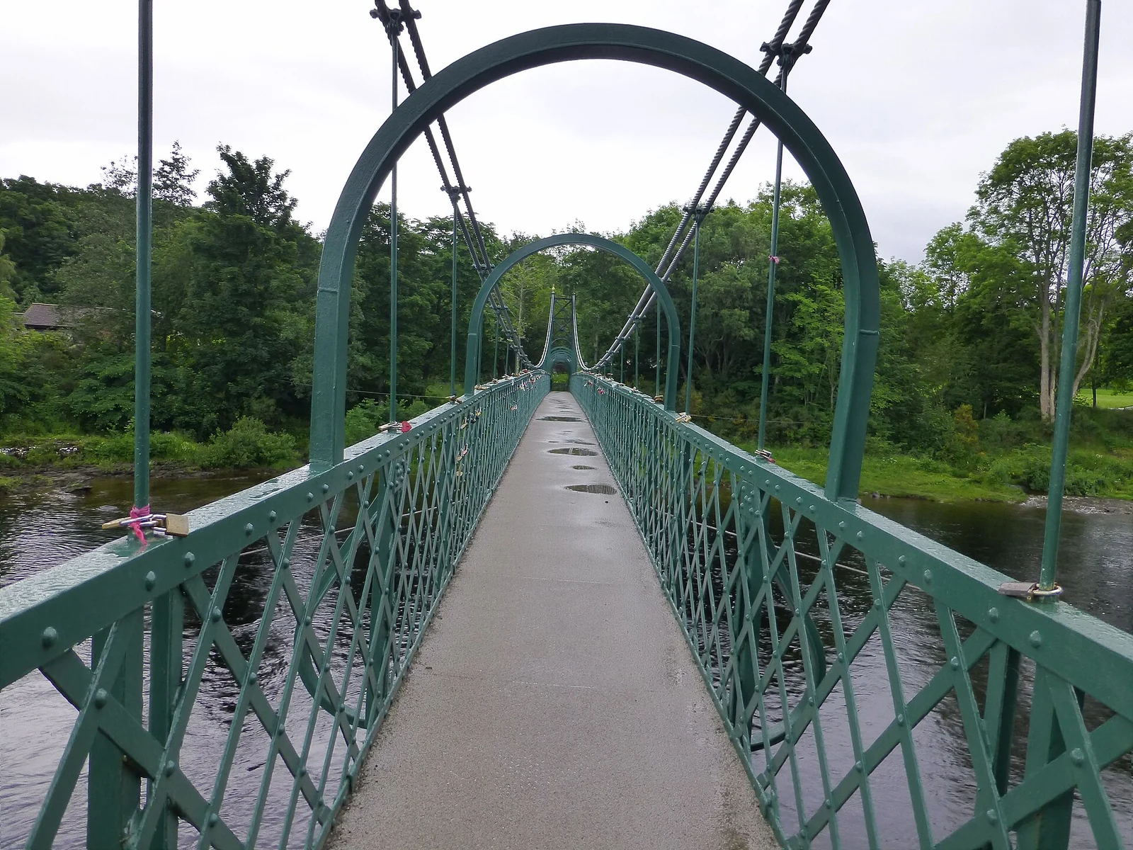 Een groene metalen voetgangershangbrug bij Pitlochry, met het pad tussen de kabels naar bomen aan de overkant.