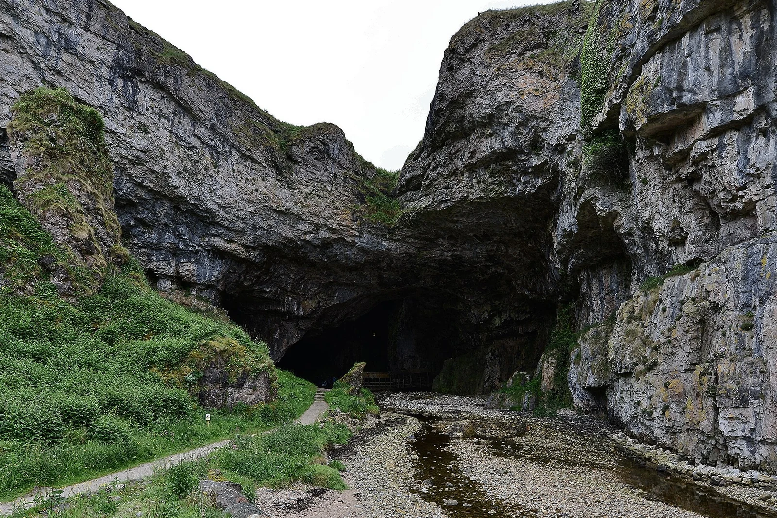De donkere ingang van Smoo Cave tussen hoge rotswanden, met een beekje en een voetpad op de voorgrond.